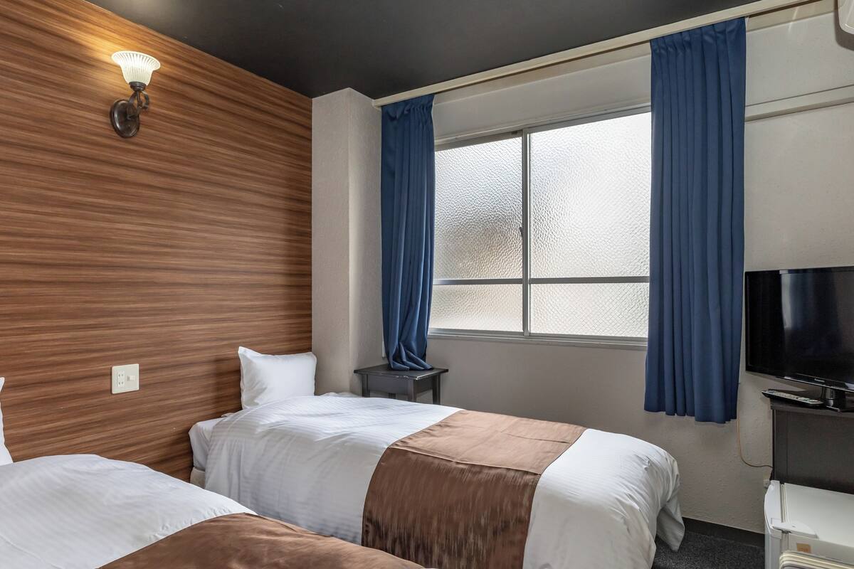 Ein Hotelzimmer mit zwei Betten, einem Fernseher, einem kleinen Tisch und einem Fenster mit Vorhängen.