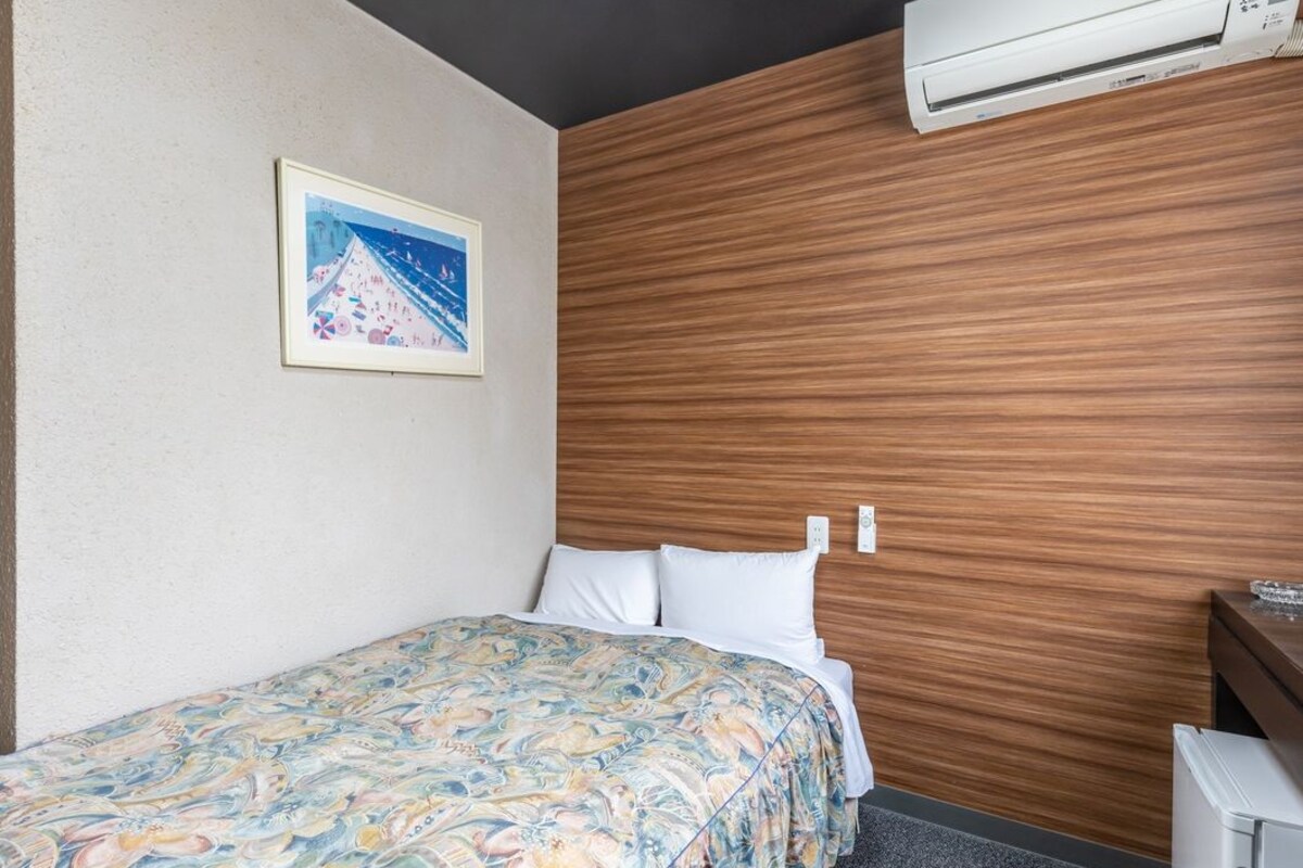 A hotel room with a bed, a wooden headboard, a framed picture on the wall, and an air conditioning unit.