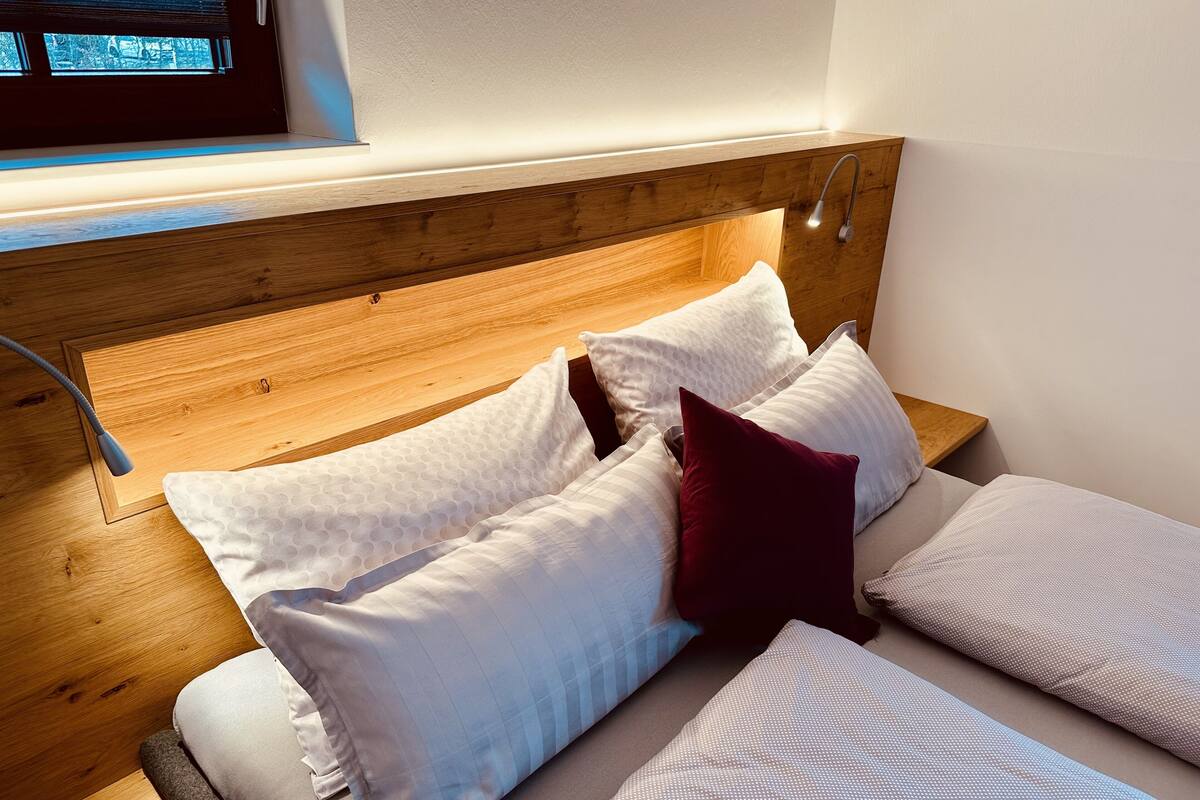 A neatly made bed with white and patterned bedding, a wooden headboard, and a window with blinds.