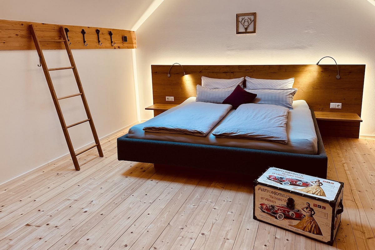 A bedroom with a wooden floor, a bed with a headboard, a ladder, and a box on the floor.