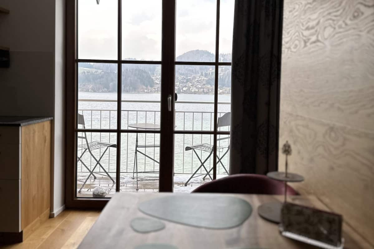 A room with a large sliding glass door leading to a balcony with a view of a body of water and hills. There is a wooden table with a glass top and a small decorative item on it.