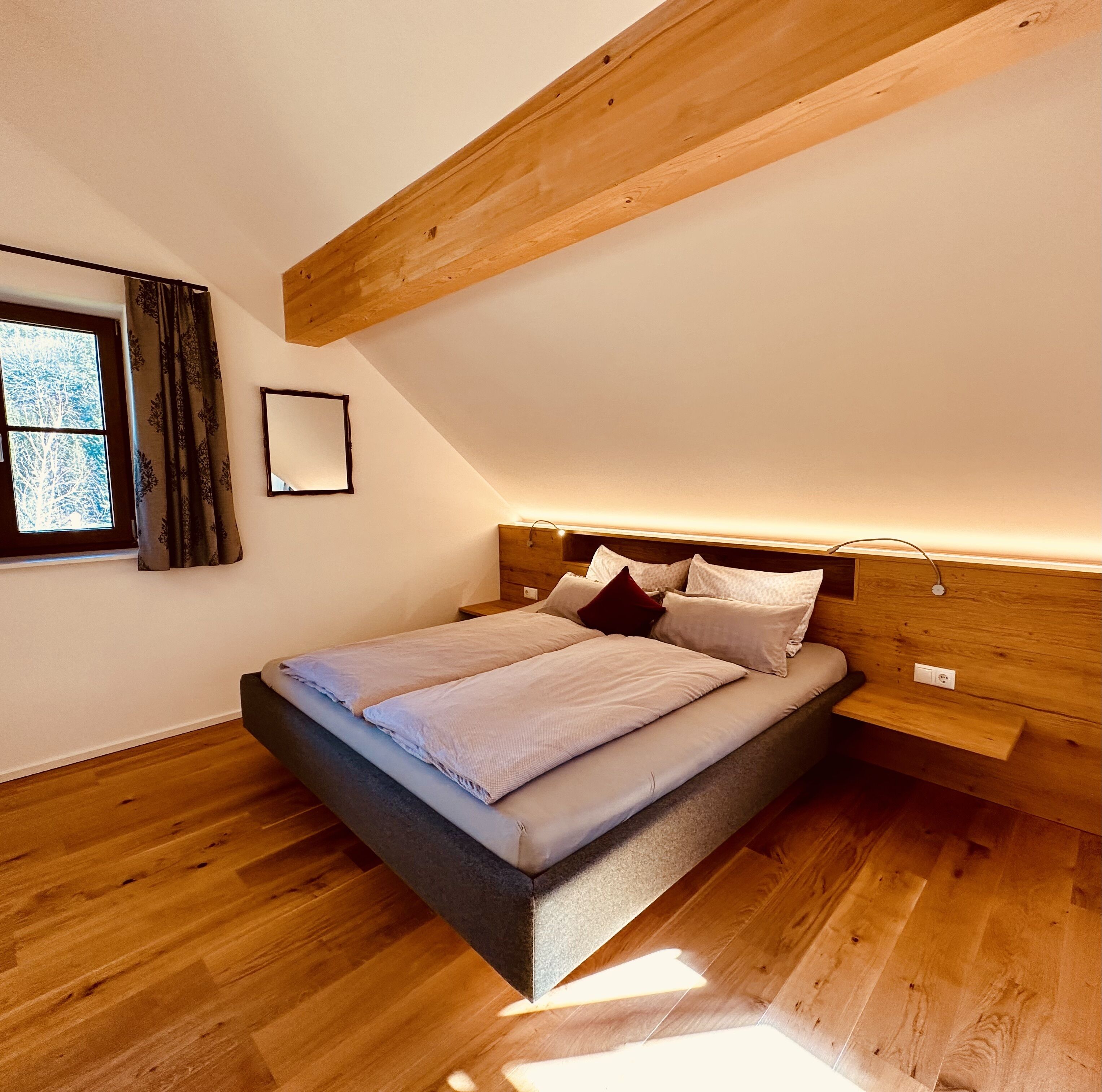Ein modernes Schlafzimmer mit schräg verlaufender Decke, einem Bett mit vielen Kissen, einem Nachttisch und einem Fenster mit Vorhängen.