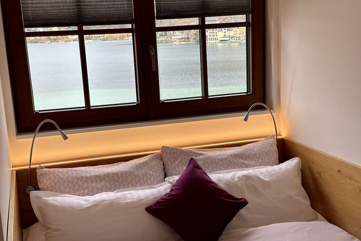 A bed with a headboard, a window with blinds, and a view of a body of water and buildings.