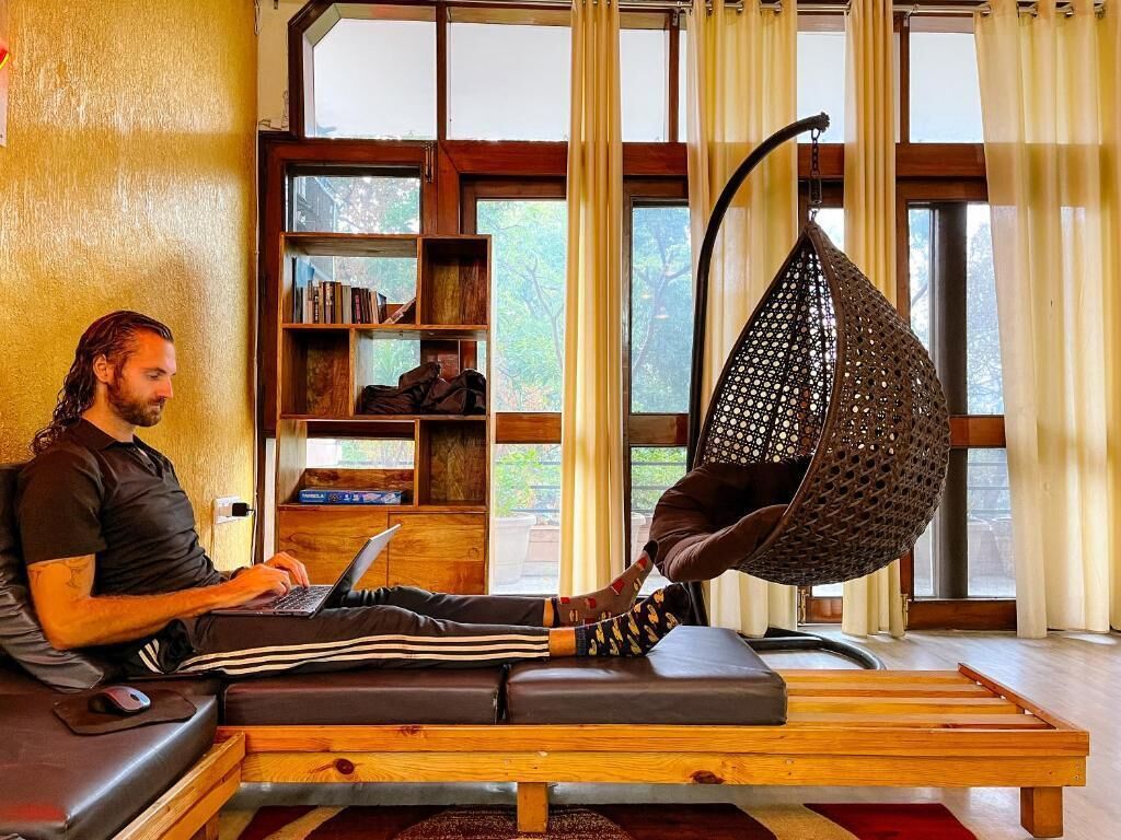 A person using a laptop on a wooden bench with a hanging chair in a room with large windows and bookshelves.
