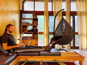A person using a laptop on a wooden bench with a hanging chair in a room with large windows and bookshelves. - Hide-In Hostel Delhi (New Delhi)