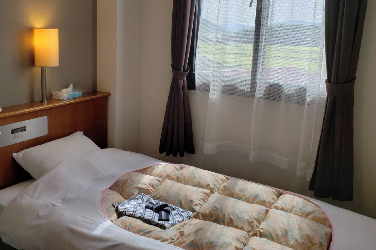 Quarto de hotel com uma cama, abajur, telefone e janela com cortinas.