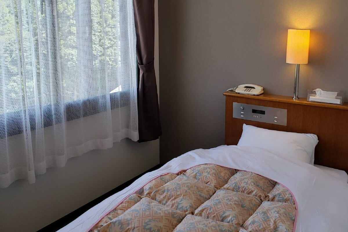 A hotel room with a bed, a bedside telephone, a lamp, and a window with sheer curtains.