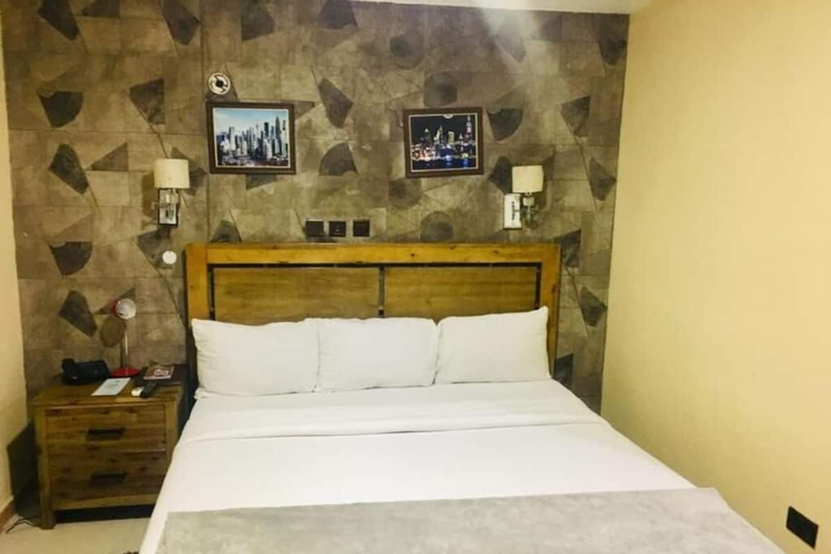 A hotel room with a bed, wooden headboard, two wall-mounted lamps, and a cityscape picture on the wall.