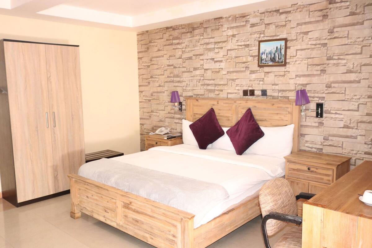 A bedroom with a wooden bed, a nightstand, a desk, a wardrobe, and a stone wall with a picture.