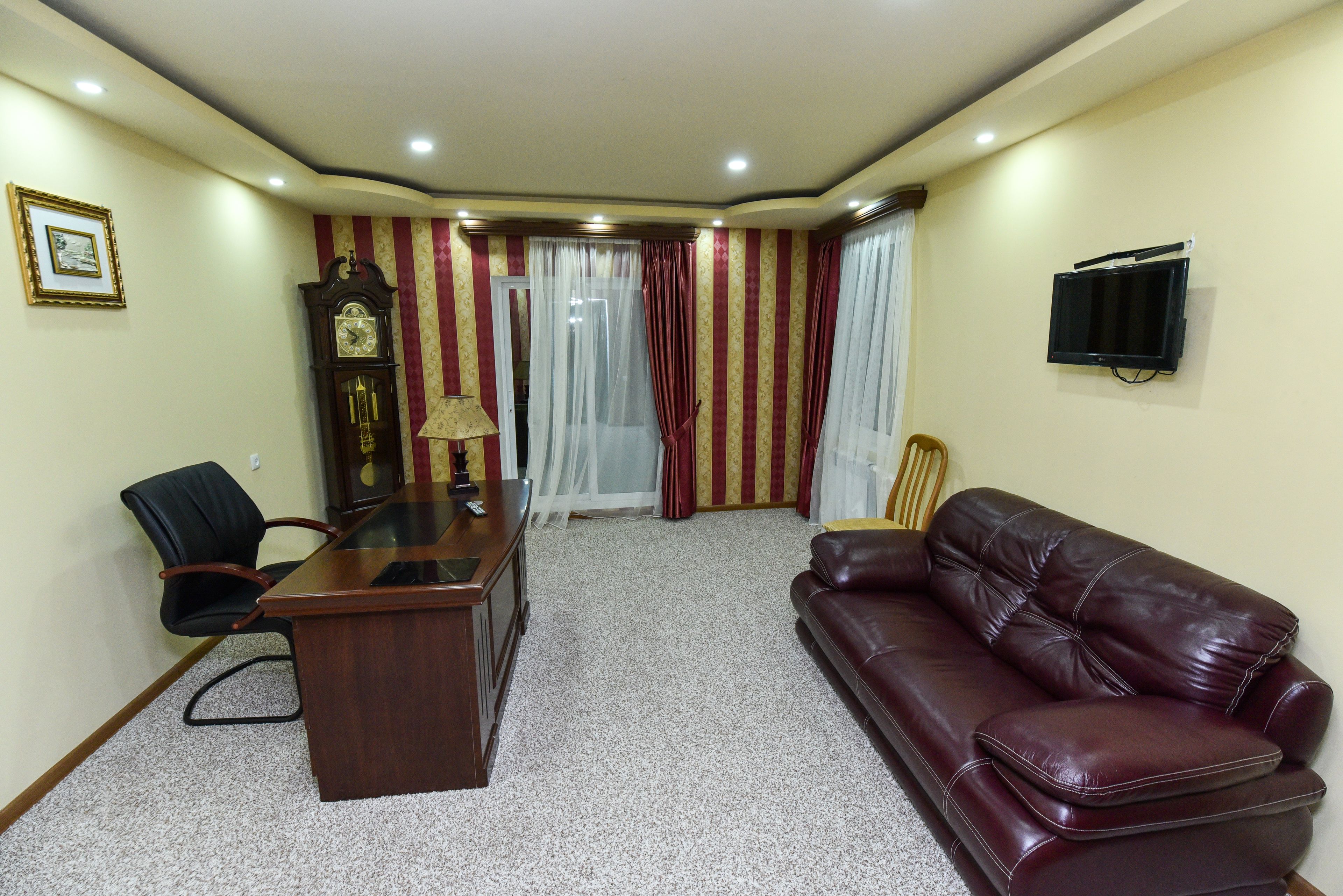 Uma sala de estar com um sofá de couro marrom escuro, uma escrivaninha de madeira com abajur, uma cadeira preta, um relógio de parede grande e uma TV fixada na parede.