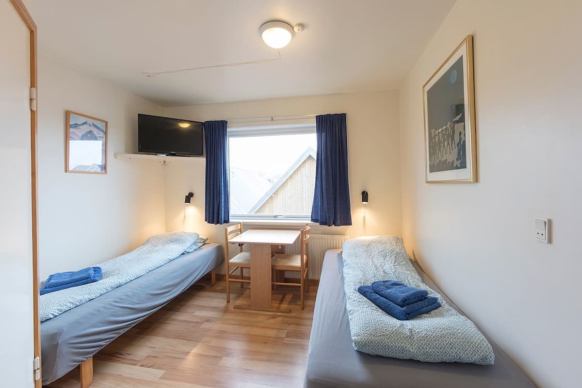 A small, cozy room with two single beds, a desk, and a chair. There is a window with curtains, a mounted TV, and a framed picture on the wall.