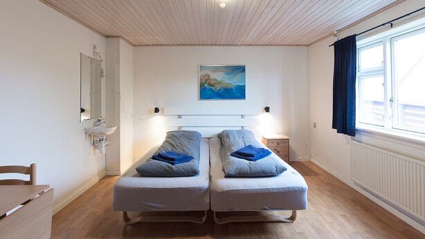 Ein Zimmer mit zwei Einzelbetten, Holzboden, weißer Decke, einem Bild an der Wand und einem Fenster mit Vorhängen.