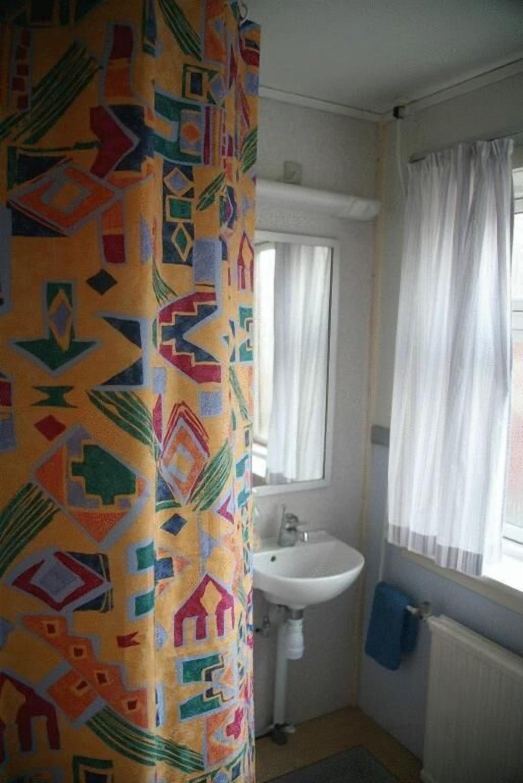 Une salle de bain avec un rideau de douche à motifs, un lavabo blanc, un miroir et une fenêtre avec des rideaux.
