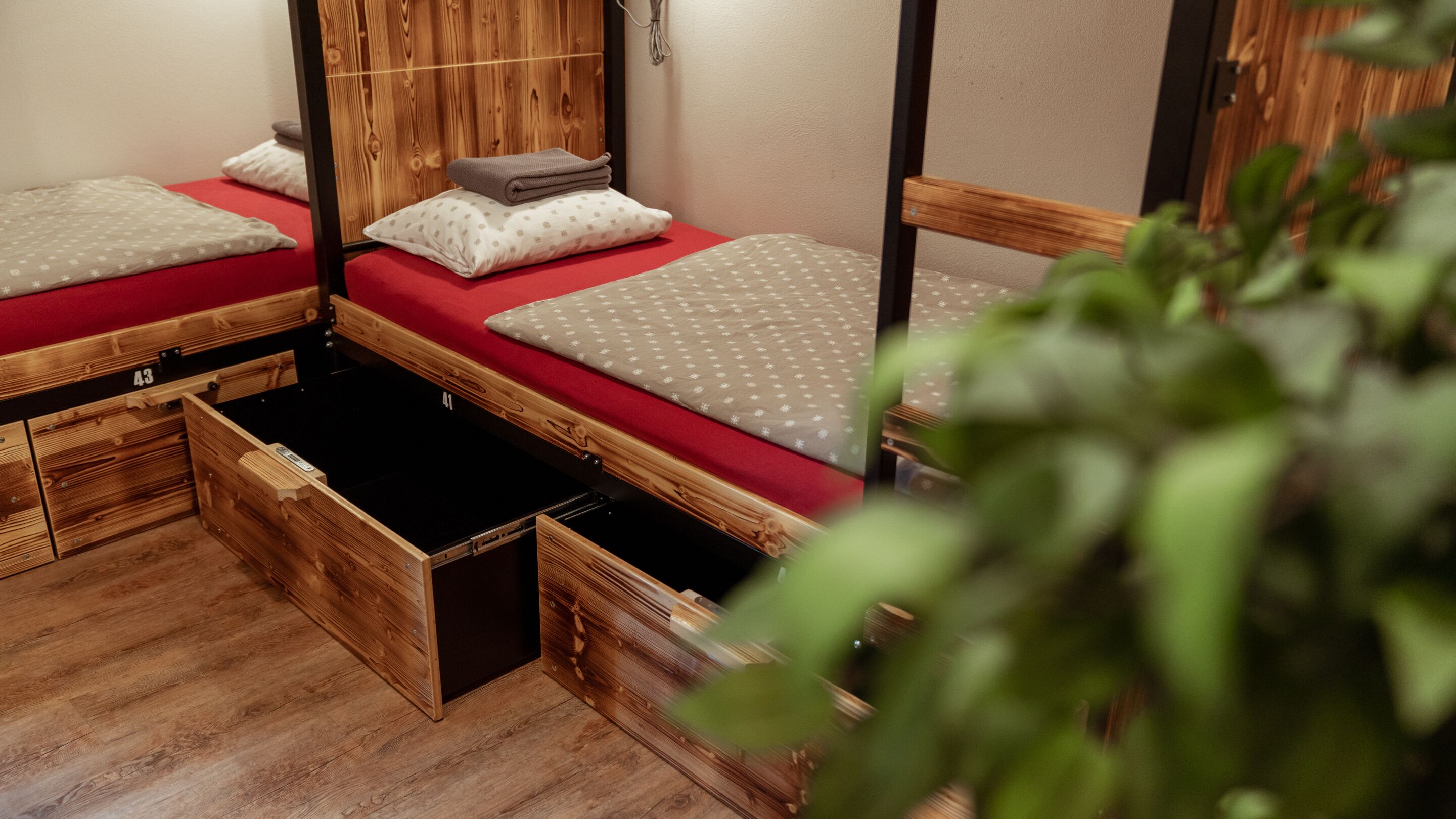 Una habitación con tres literas de madera, cada una con colchón rojo y almohada. Hay cajones de madera debajo de las camas, y una planta se ve parcialmente al frente.