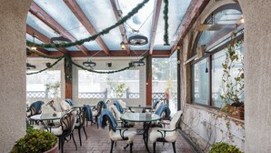 Un patio al aire libre con mesas y sillas redondas, cubierto por un techo de vidrio y decorado con plantas y guirnaldas luminosas.