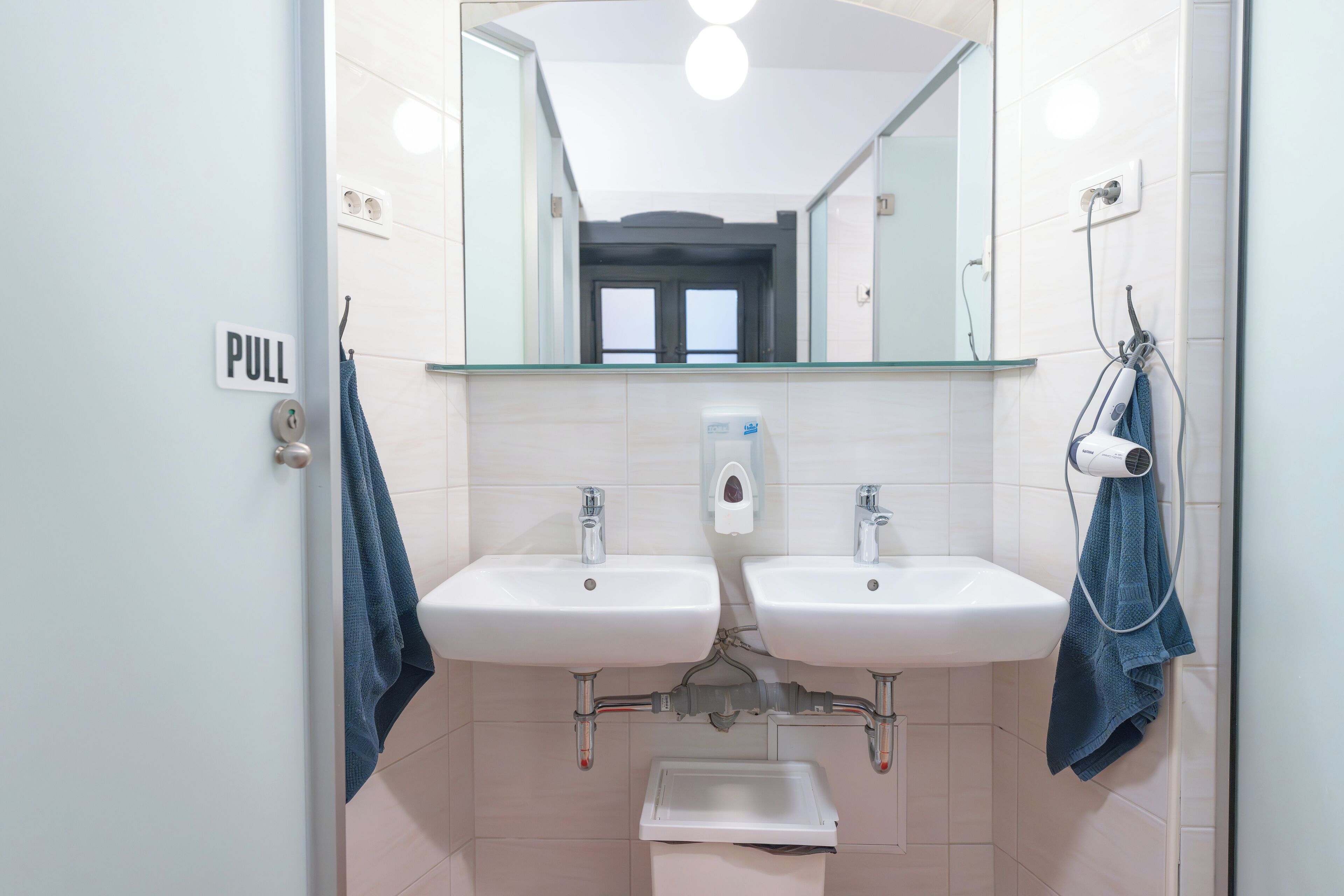 A bathroom with two sinks, a mirror, a towel rack, and a soap dispenser.