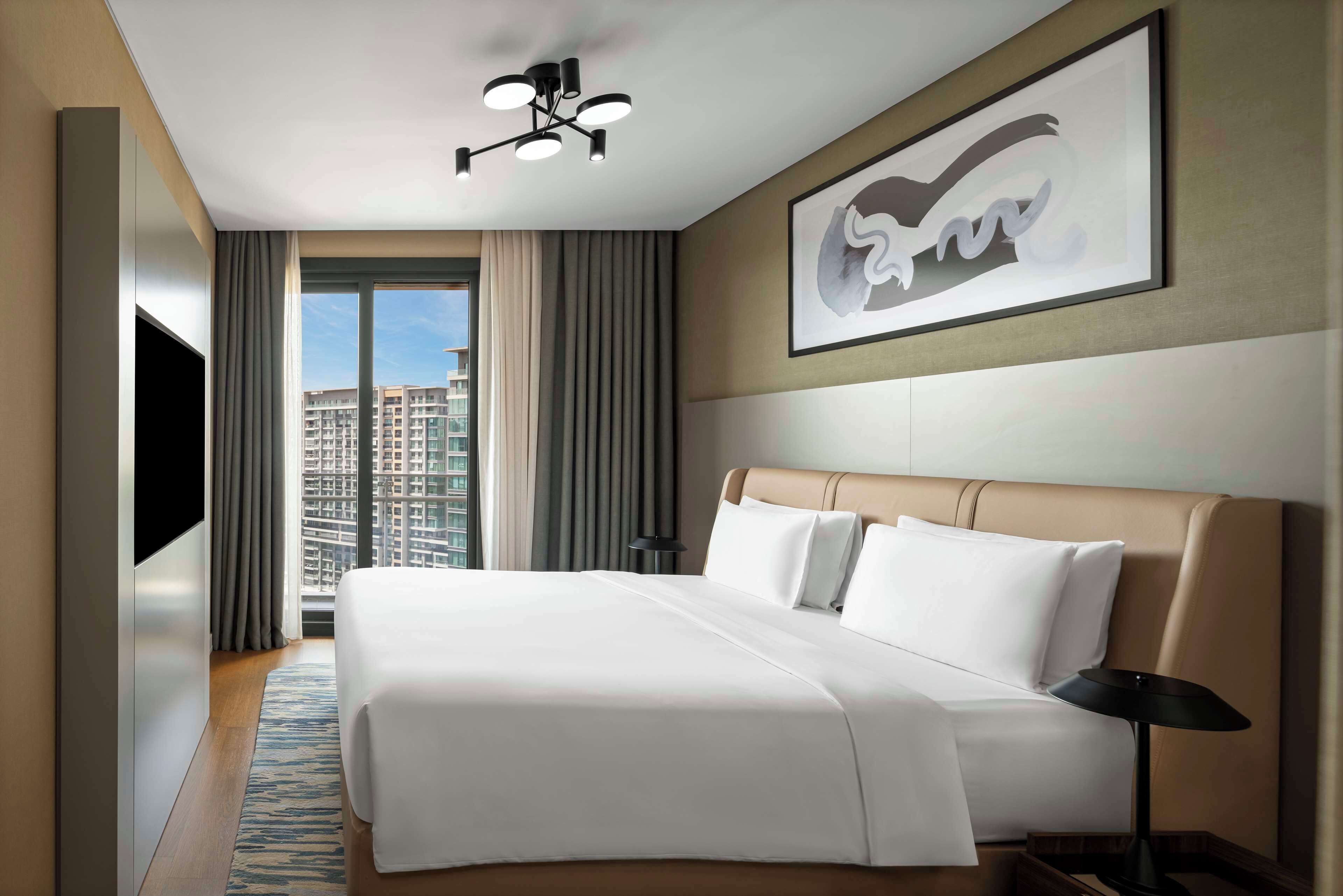 A hotel room with a large bed, a flat-screen TV, a ceiling light, a framed artwork, and a view of a cityscape through the window.