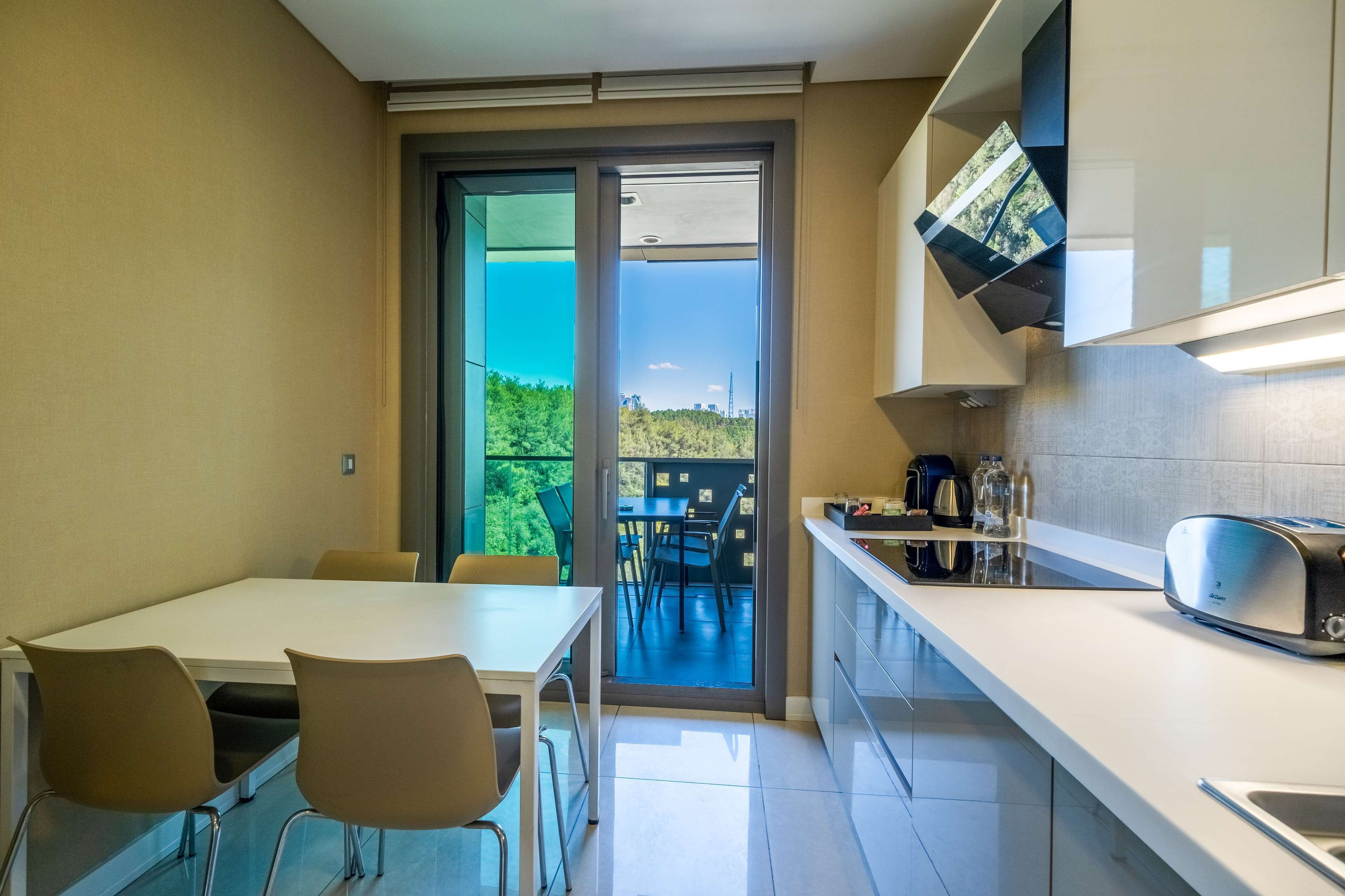 Una cocina moderna con comedor, un amplio ventanal corredera que da a un balcón exterior, y vistas a zonas verdes y edificios.