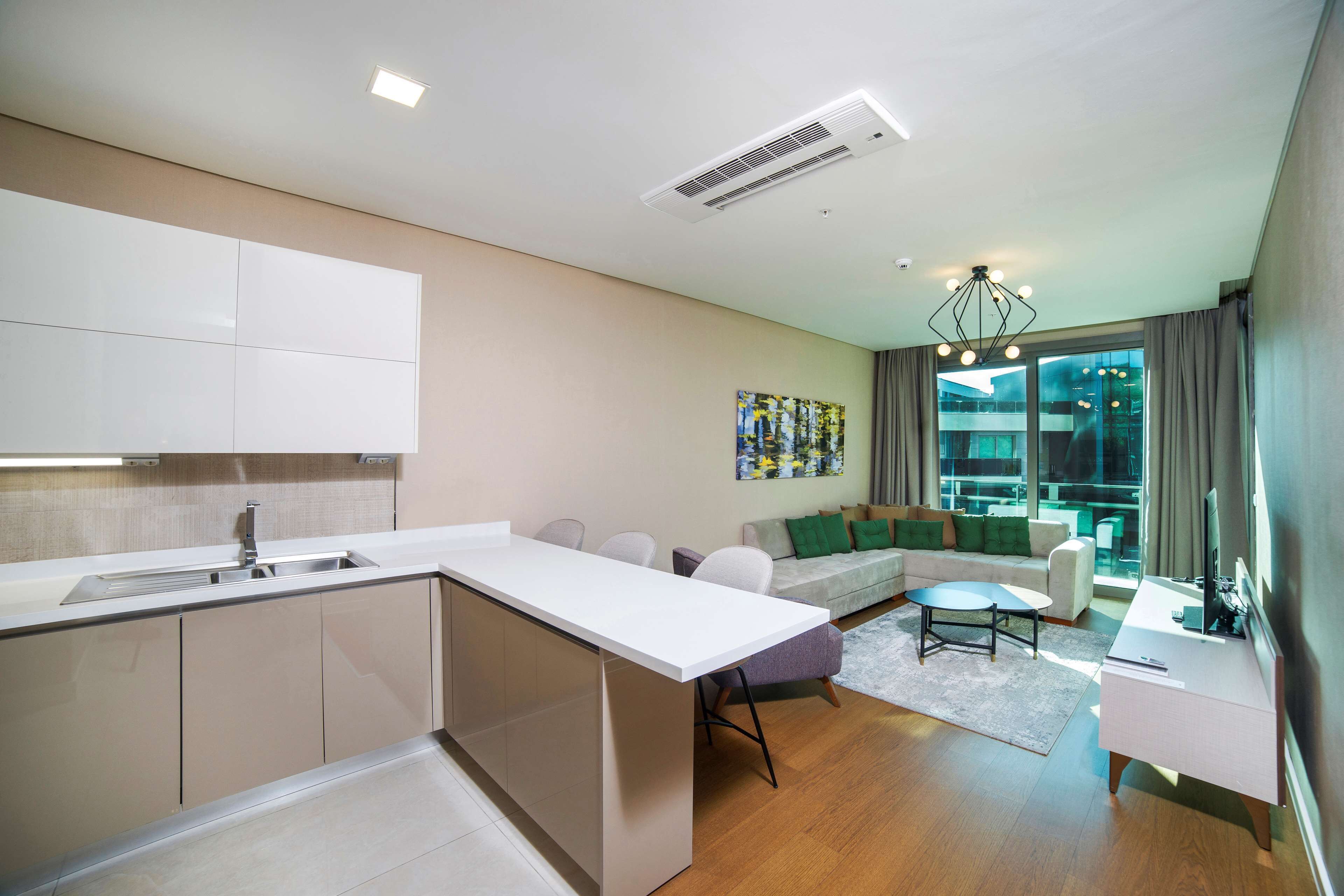 A modern kitchen with a built-in island, a dining area with a small table and chairs, a sofa, and a large window offering a view of the city.