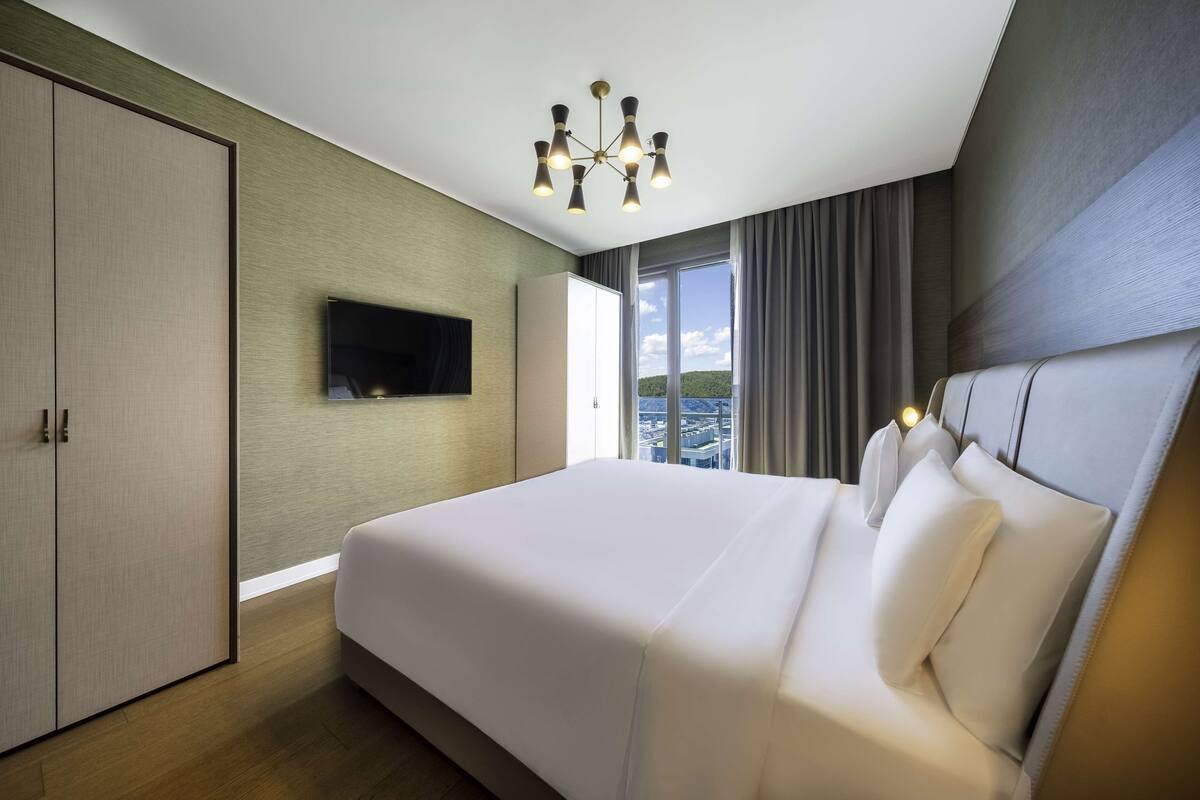 A modern hotel room with a large bed, a flat-screen TV, a wardrobe, and a view of the city through large windows.
