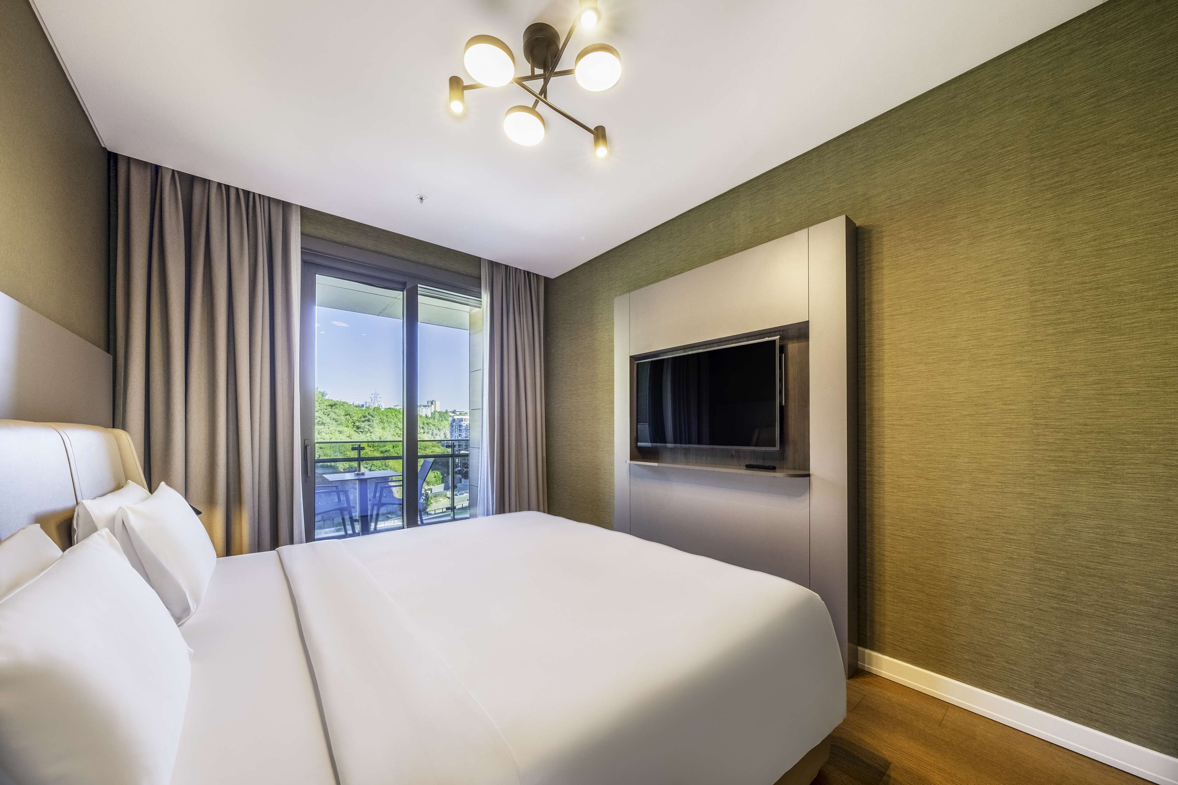 A hotel room with a large bed, a TV mounted on the wall, and a balcony with a view of greenery.