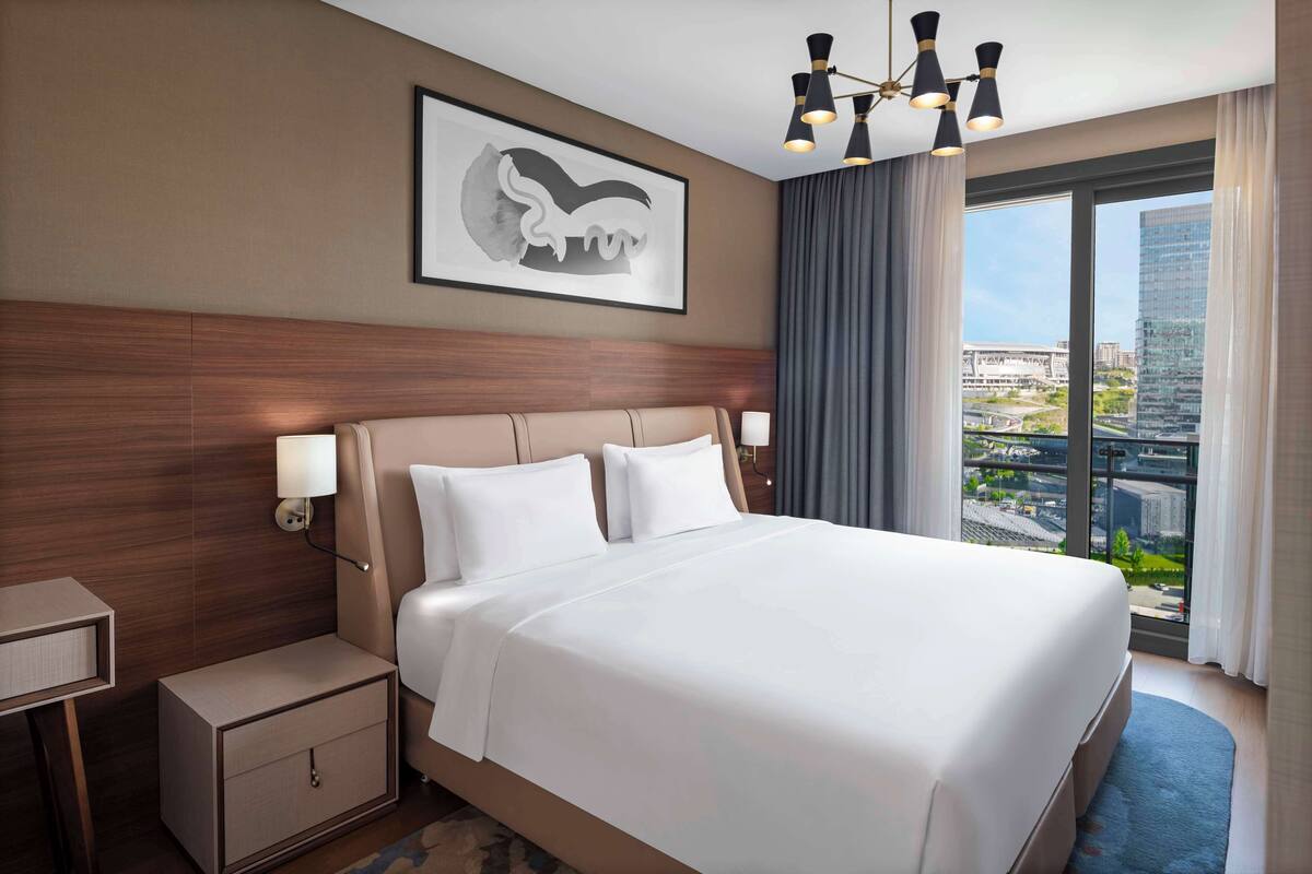 A hotel room with a large bed, a nightstand, a wall-mounted lamp, a framed artwork, and a view of the city through a large window.