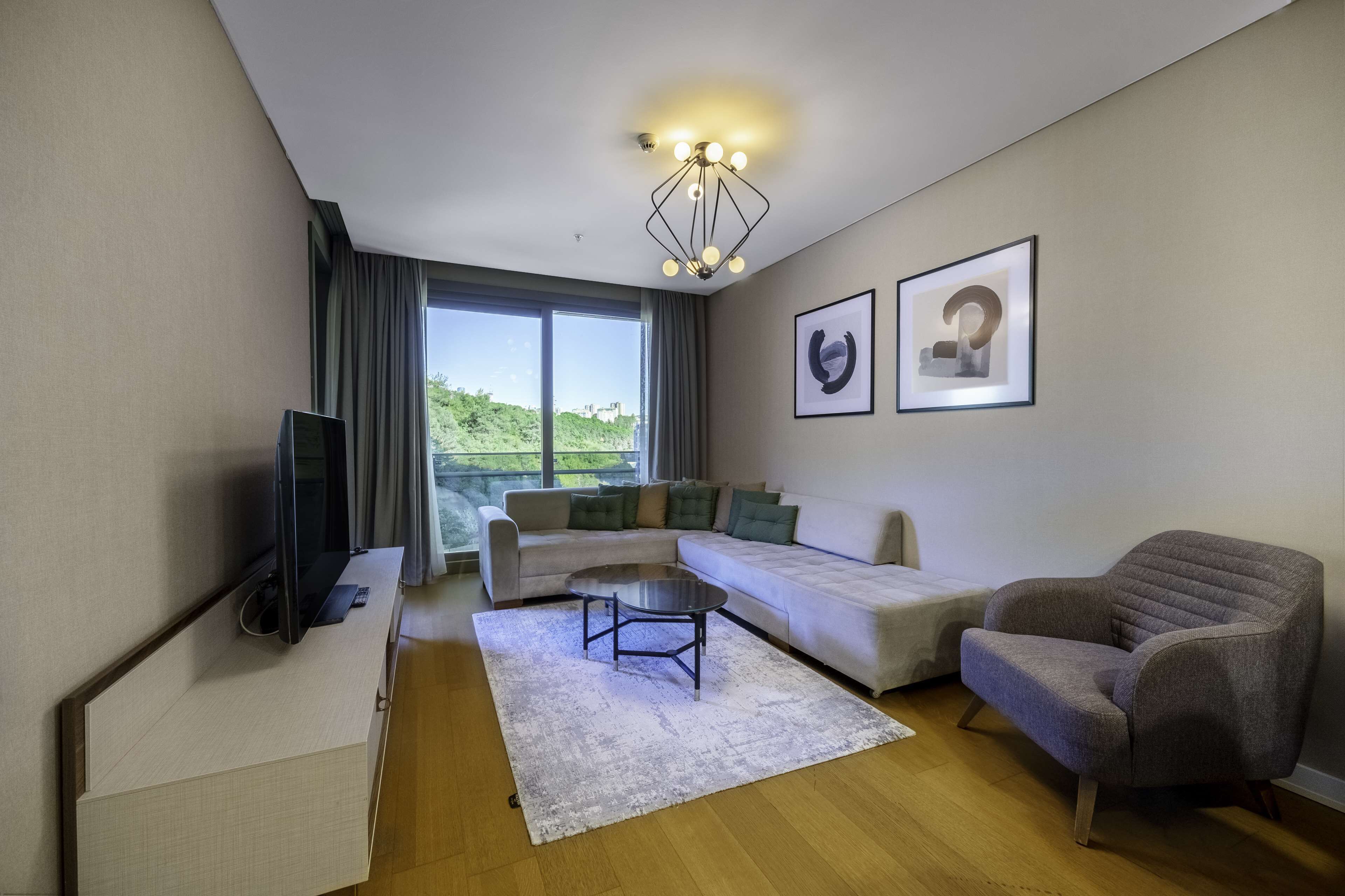 A modern living room with a sofa, armchair, and coffee table. There is a TV on a stand, a large window with curtains, and two framed pictures on the wall.