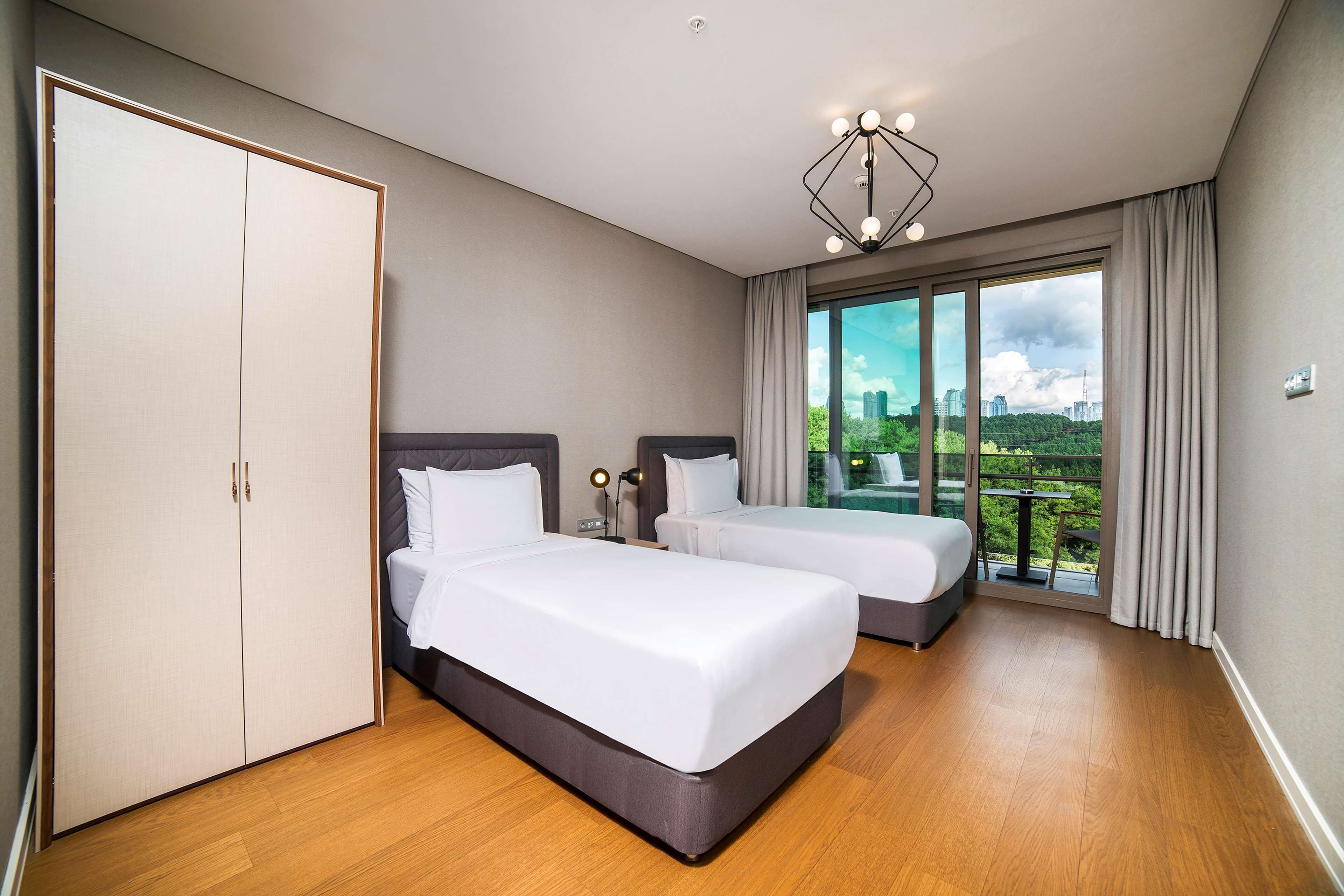 Une chambre d’hôtel avec deux lits, une armoire, un balcon donnant sur la ville et un lustre moderne.