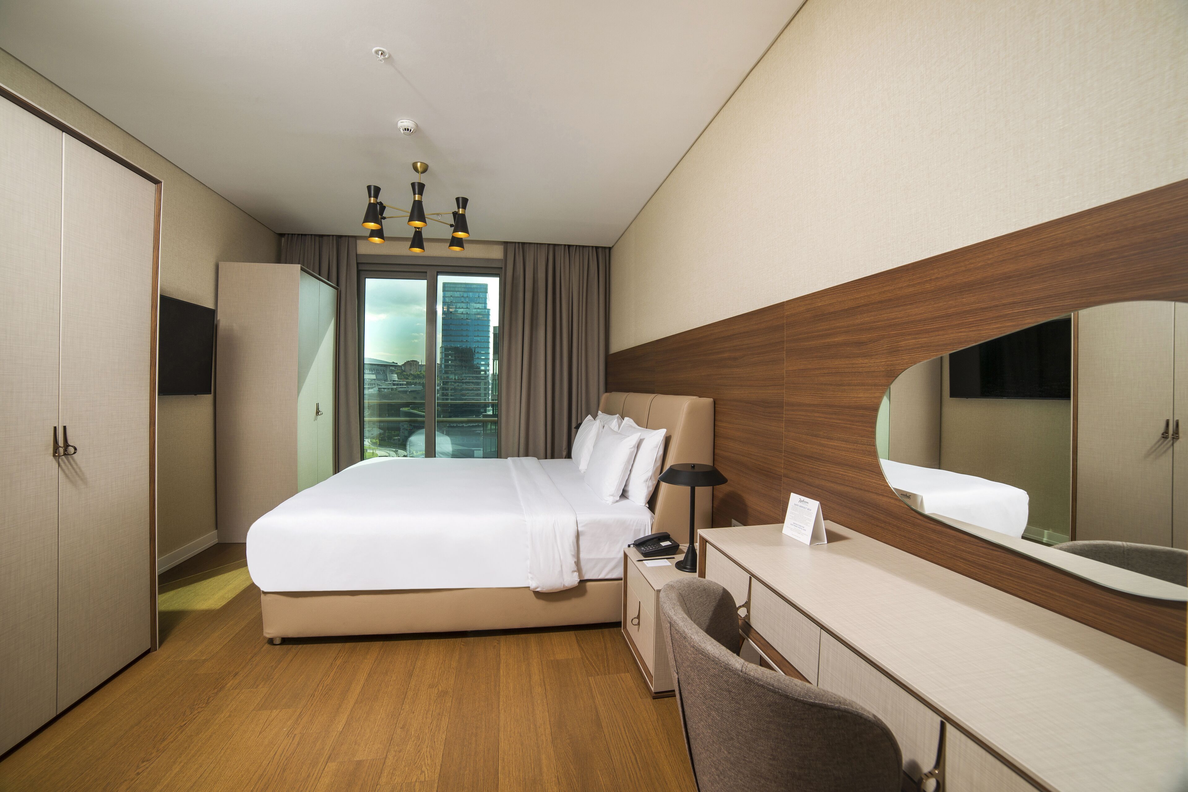 A modern hotel room with a large bed, a desk, a chair, a mirror, and a wardrobe.