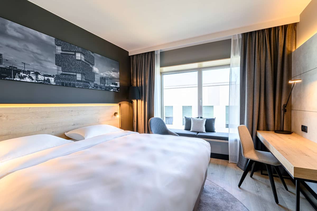 A hotel room with a large bed, a desk, a chair, and a view of the city through a large window.