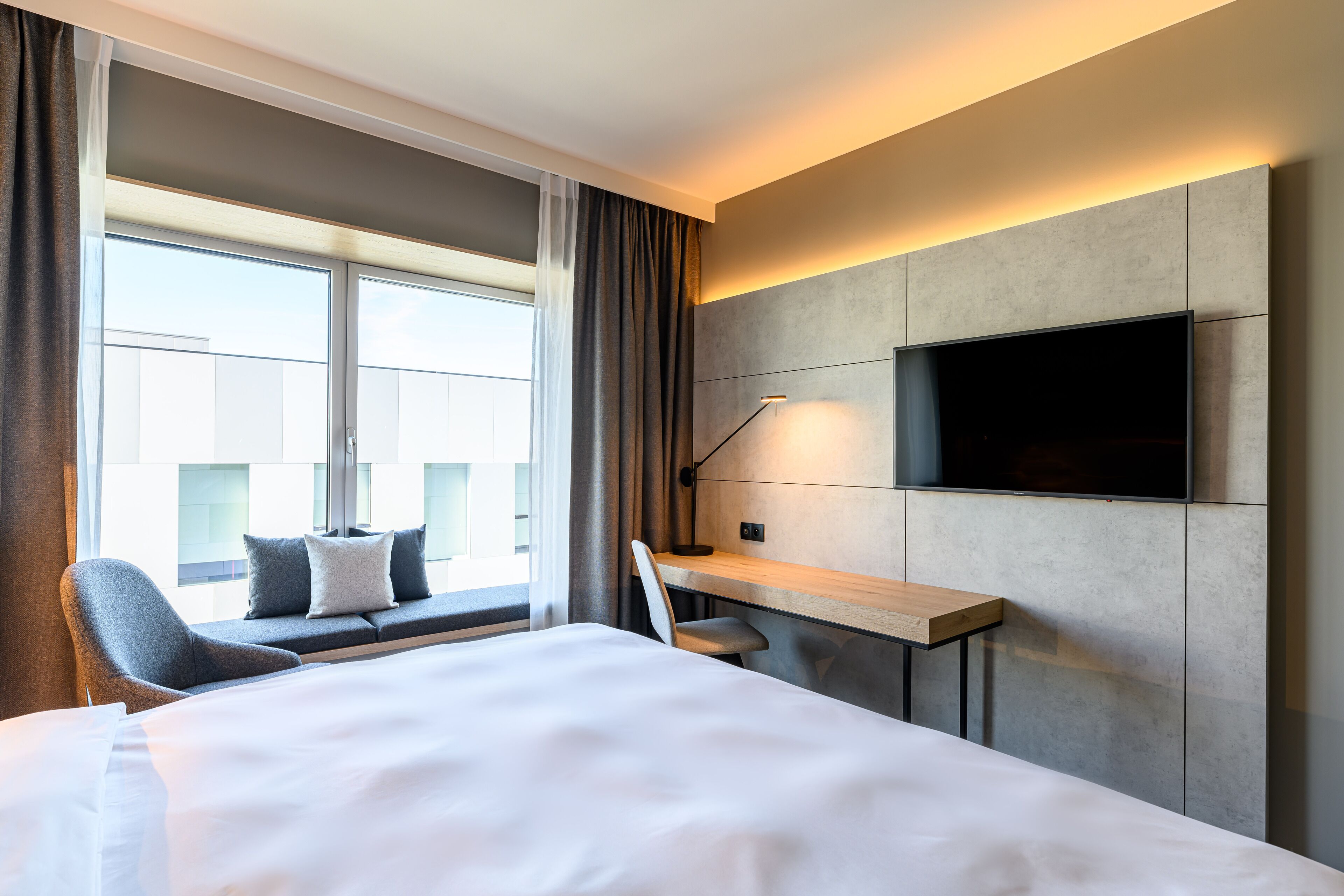 Una camera d'albergo con un letto grande, una scrivania con una sedia, una TV montata a parete e una finestra con tende.