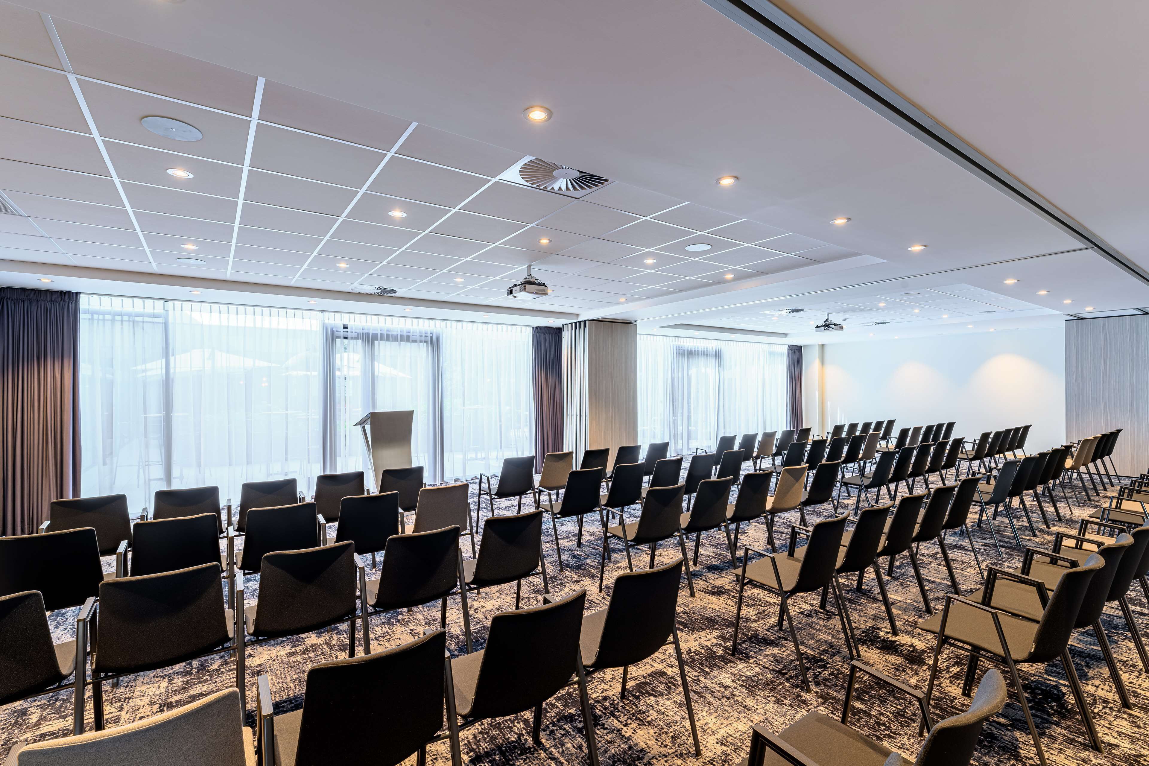 Un salón de conferencias con filas de sillas, ventanas amplias, iluminación empotrada en el techo y un proyector.