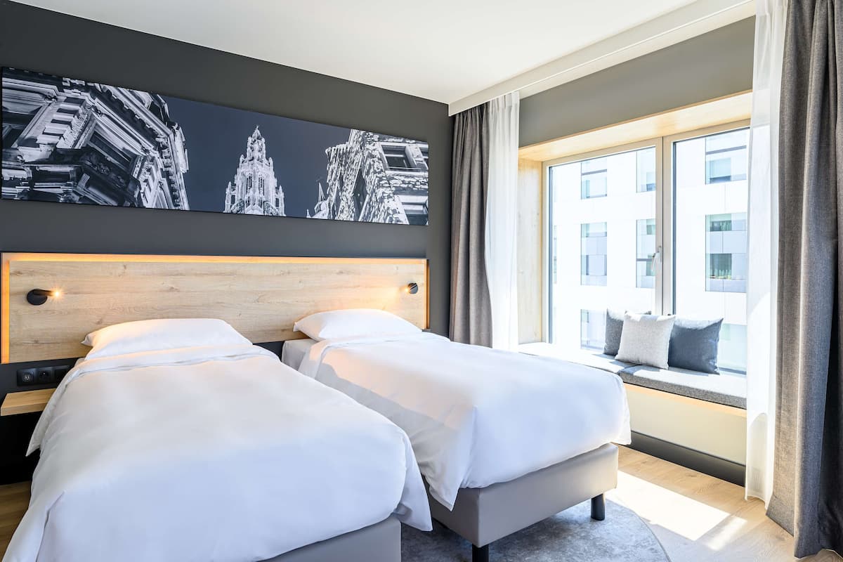 A hotel room with two beds, a large window with a view of buildings, and a wall-mounted artwork.