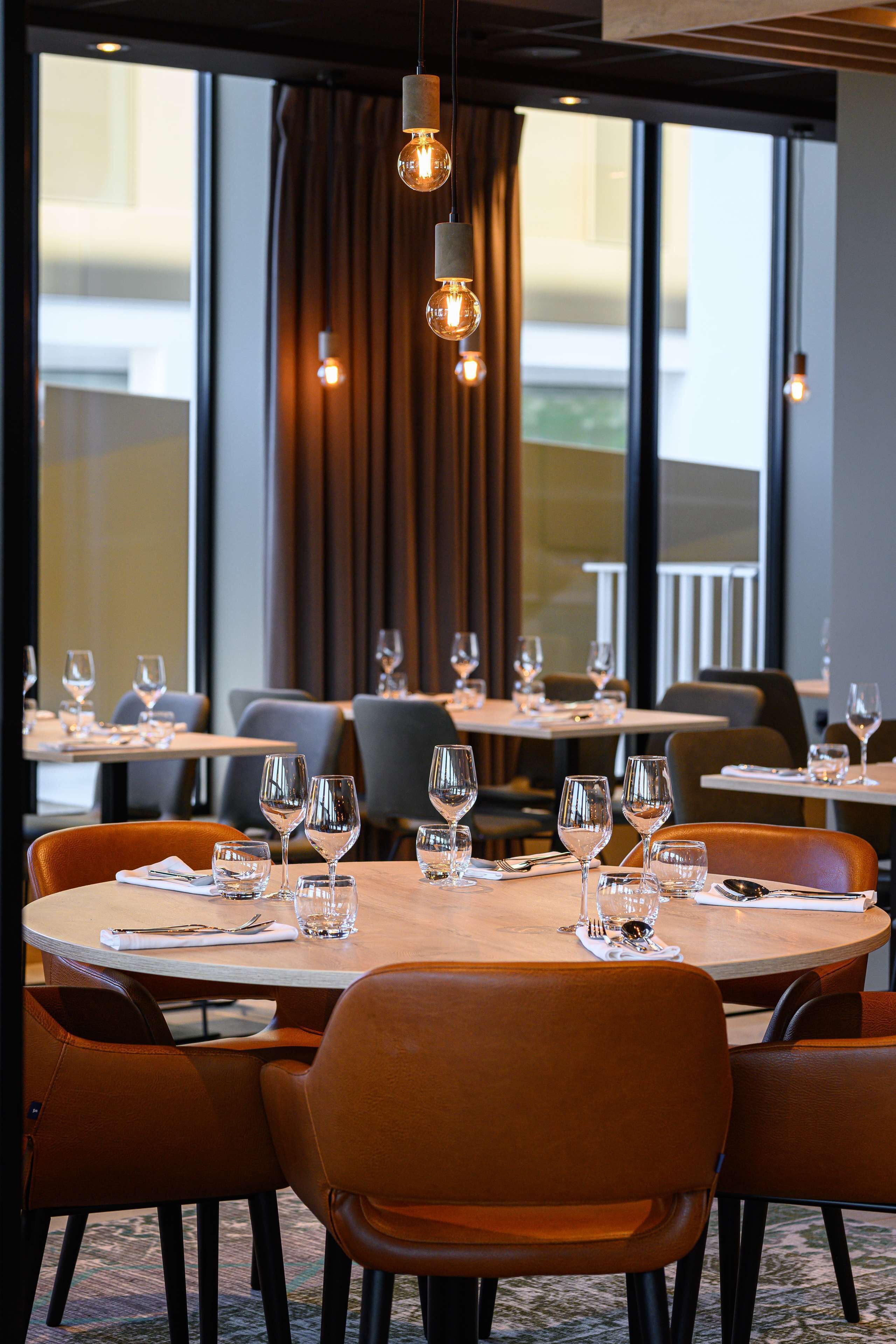 Ein Restaurant mit runden Tischen, braunen Ledersesseln und hängenden Pendelleuchten.