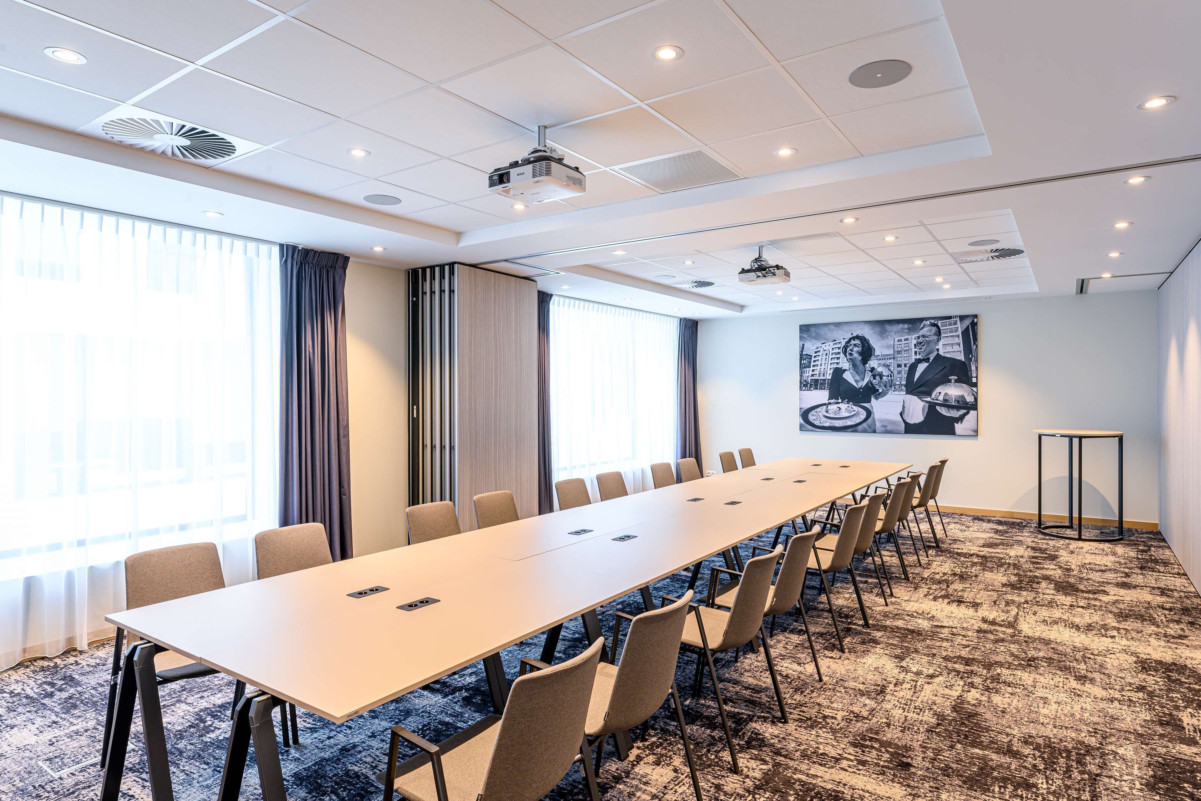 Un salón de conferencias con una larga mesa, sillas, un proyector y un amplio ventanal con cortinas.