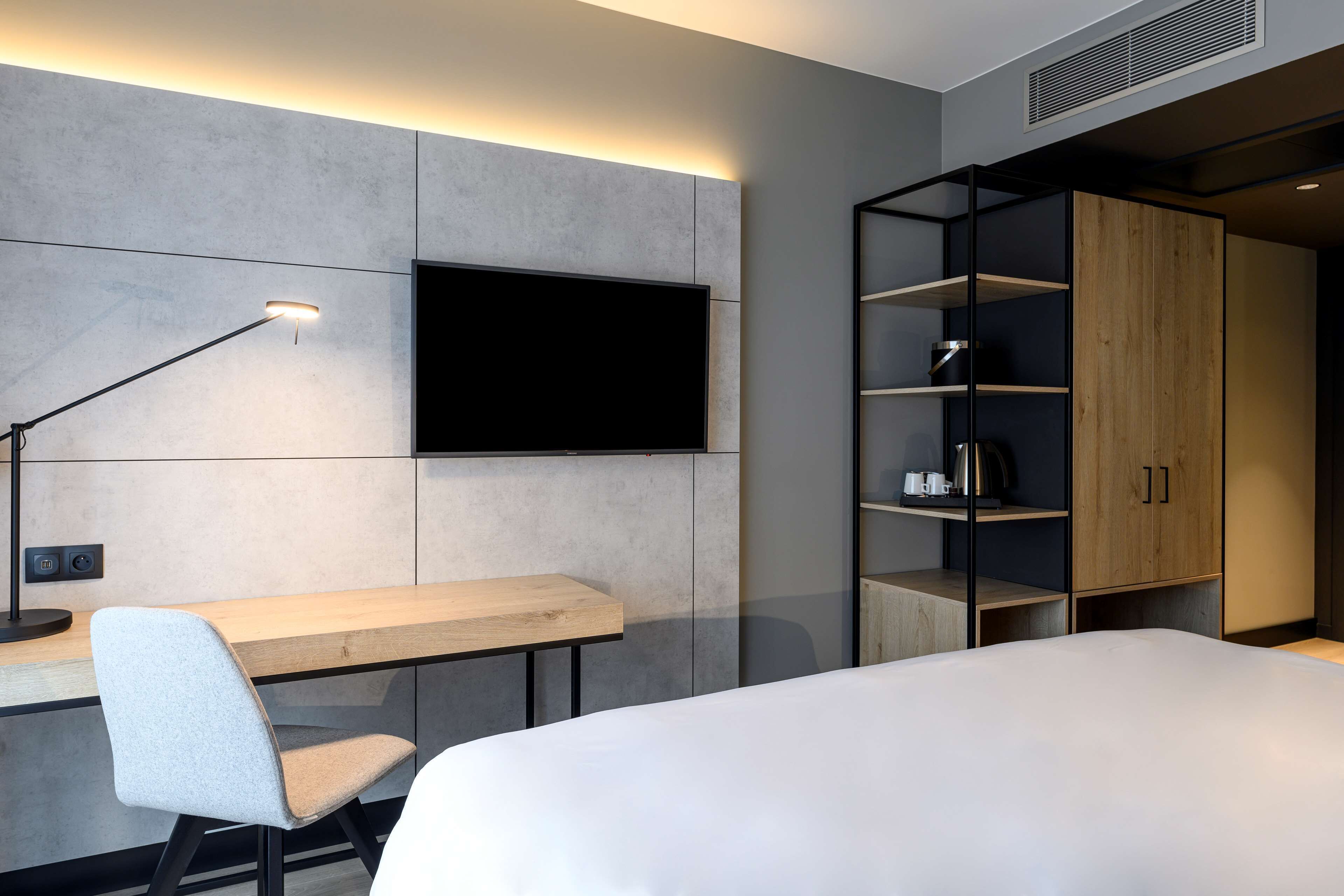 A modern hotel room with a flat-screen TV mounted on the wall, a wooden desk with a chair, a bed with white bedding, and a built-in wardrobe with shelves.