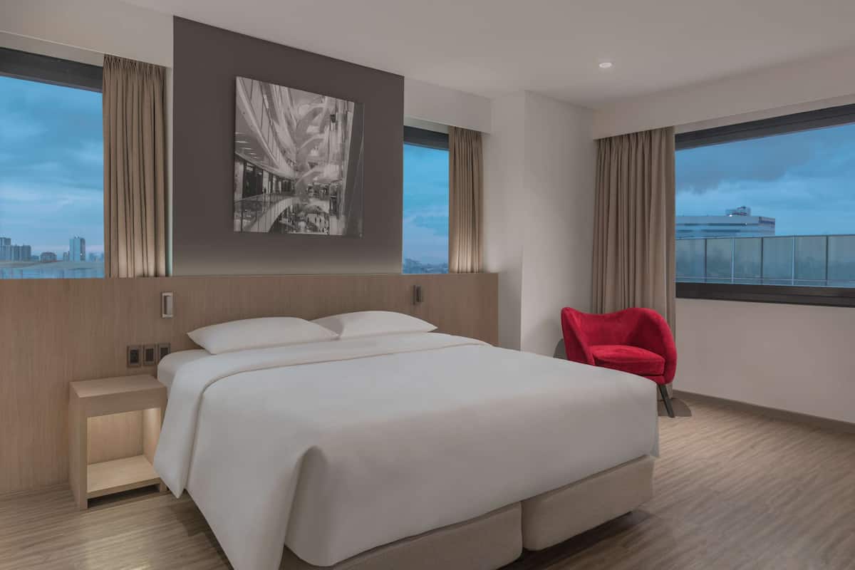 A modern hotel room with a large bed, a red chair, a city view, and a painting on the wall.