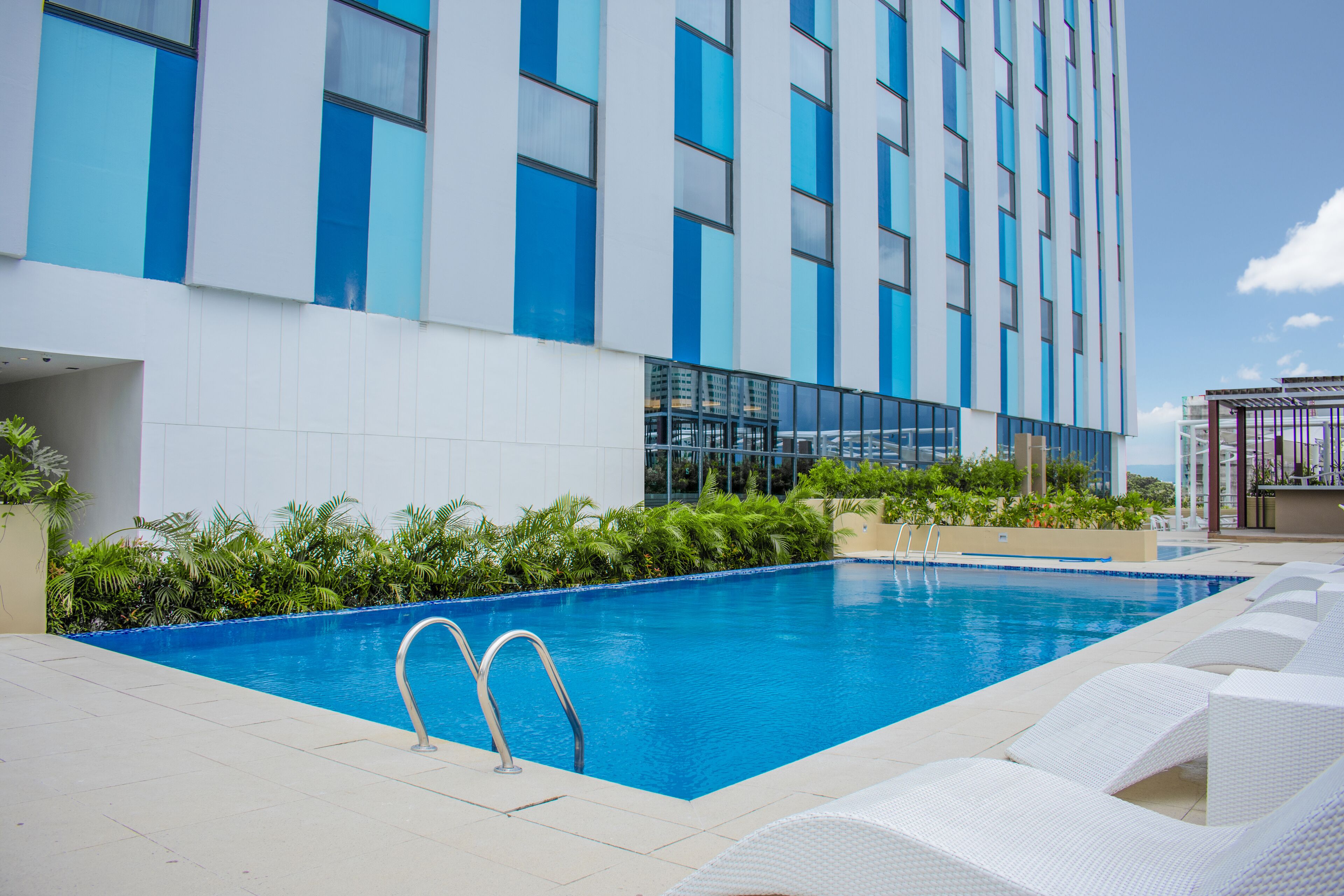 A modern hotel with a rooftop pool, white lounge chairs, and lush greenery.