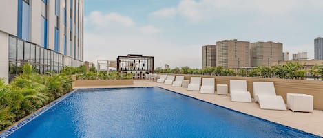 A rooftop pool with lounge chairs and a glass-enclosed area, surrounded by high-rise buildings.
