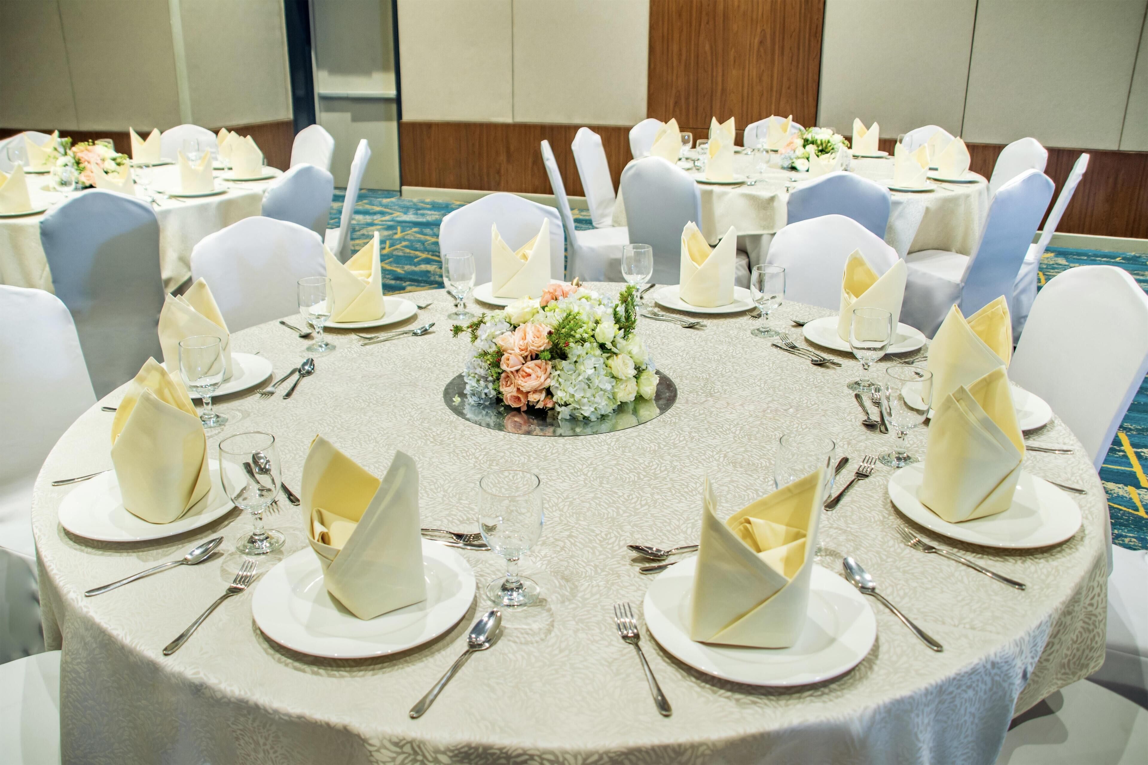 A banquet hall with round tables set for a formal event, featuring white tablecloths, neatly folded napkins, and floral centerpieces.