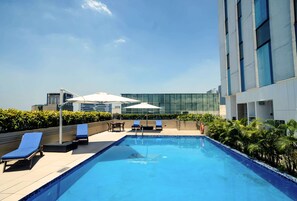 Una piscina en la azotea con sillones de descanso y sombrillas, rodeada de edificios modernos y vegetación.