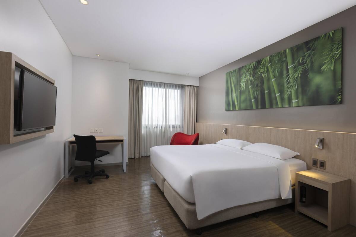 A hotel room with a bed, desk, chair, TV, and a large wall painting.