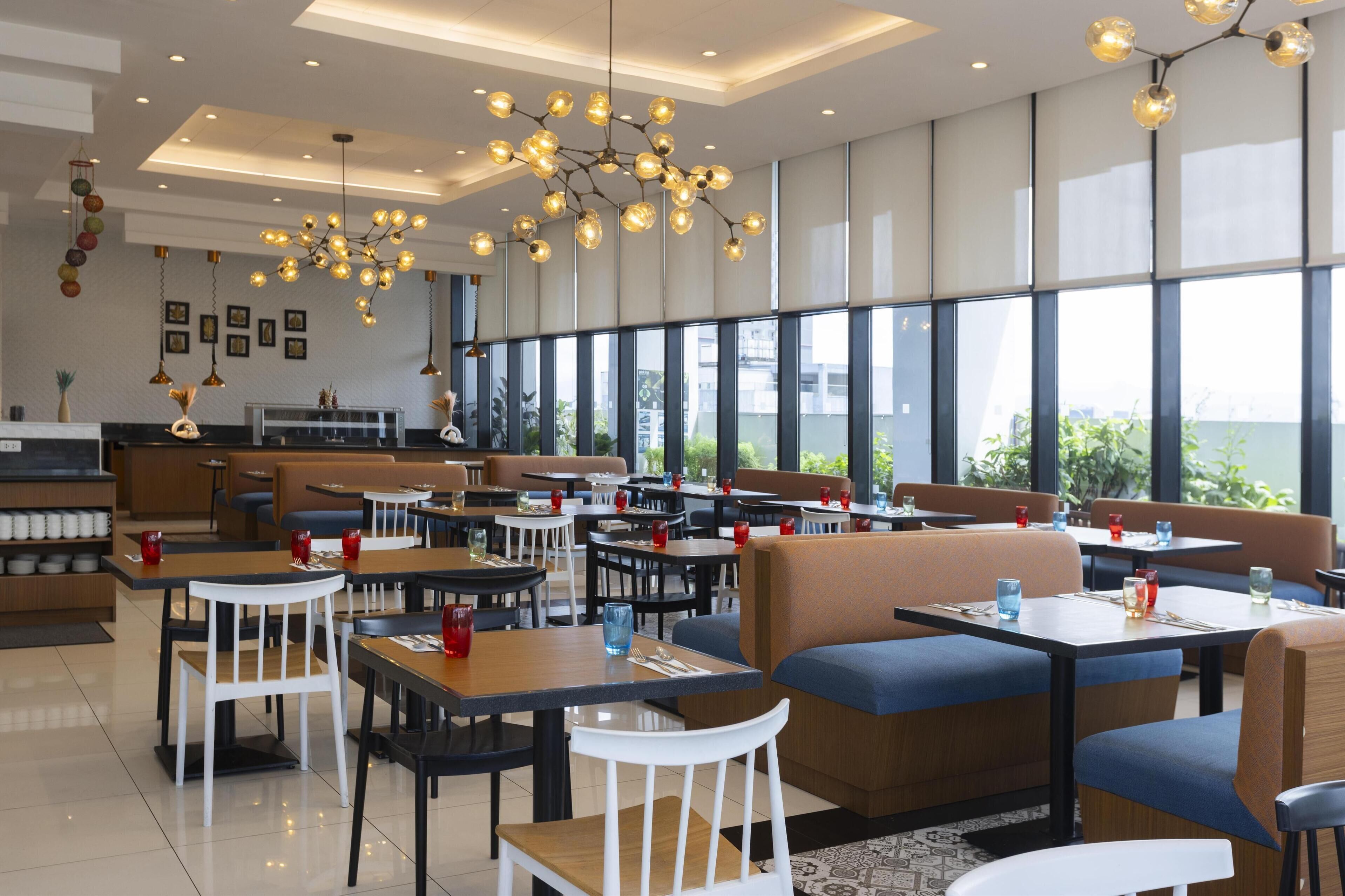 A modern restaurant with high ceilings, pendant lighting, and large windows offering a view of the outdoors.