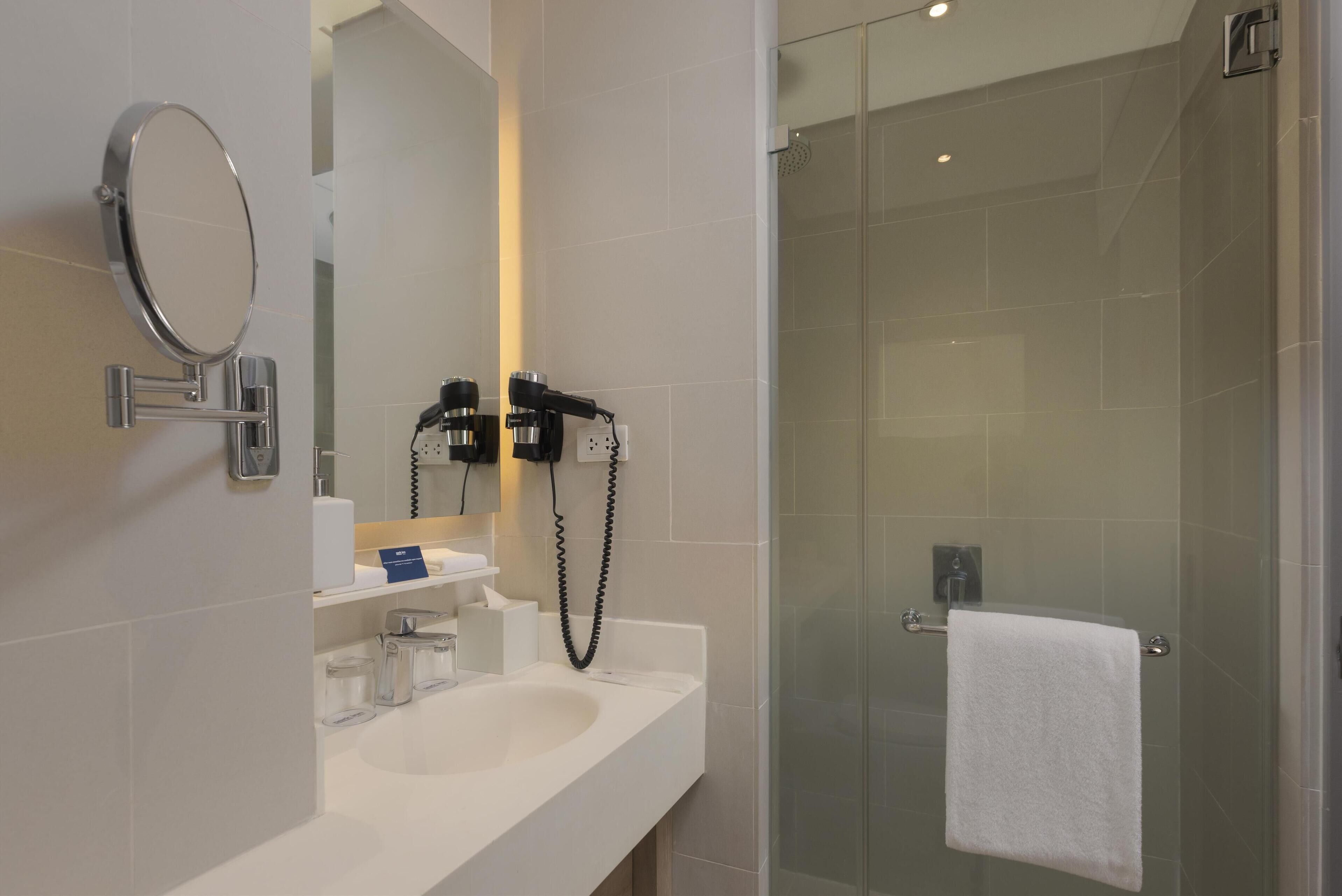 A modern bathroom with a white sink, a round mirror, a hairdryer, and a glass-enclosed shower.