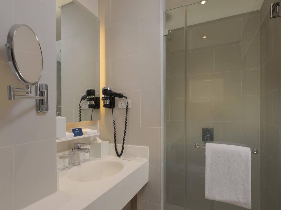 A modern bathroom with a white sink, a round mirror, a hairdryer, and a glass-enclosed shower.