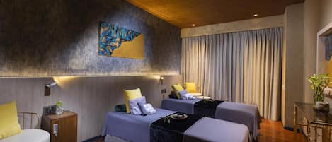 A hotel room with two beds, a small table, a chair, a bedside table with a lamp, and a large painting on the wall.