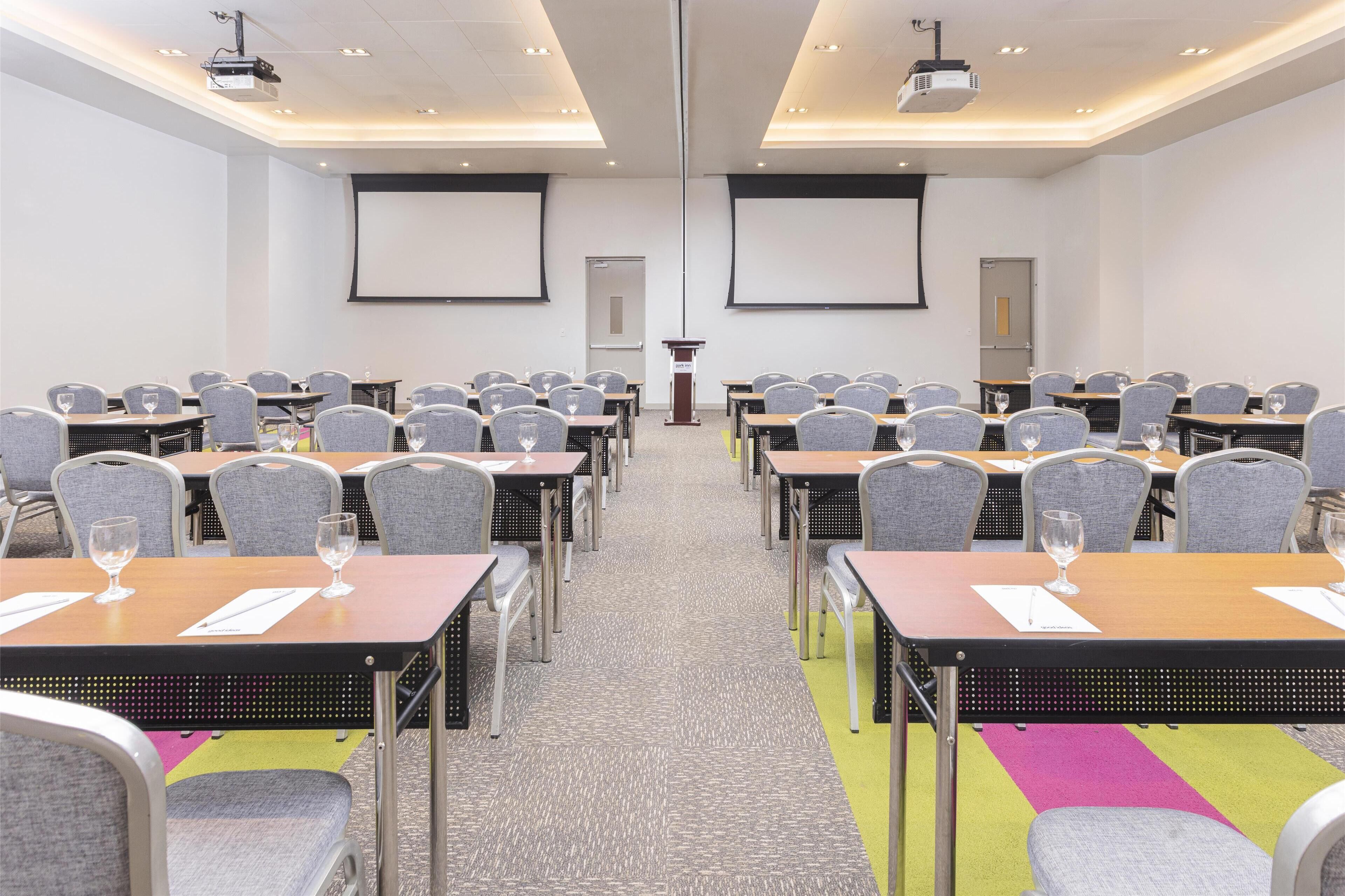 A conference room with rows of tables and chairs, a projector screen, and a podium.