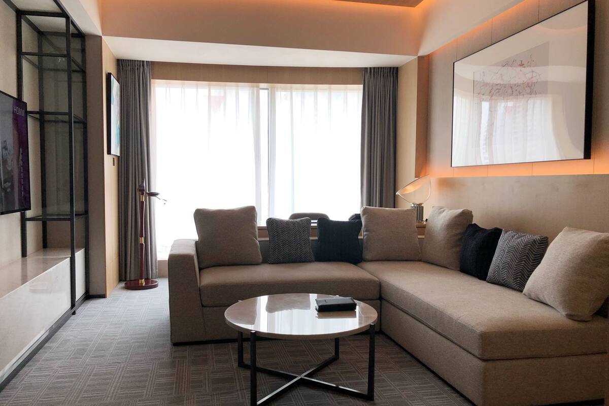 A modern living room with a beige sectional sofa, a round coffee table, and a large window with curtains.