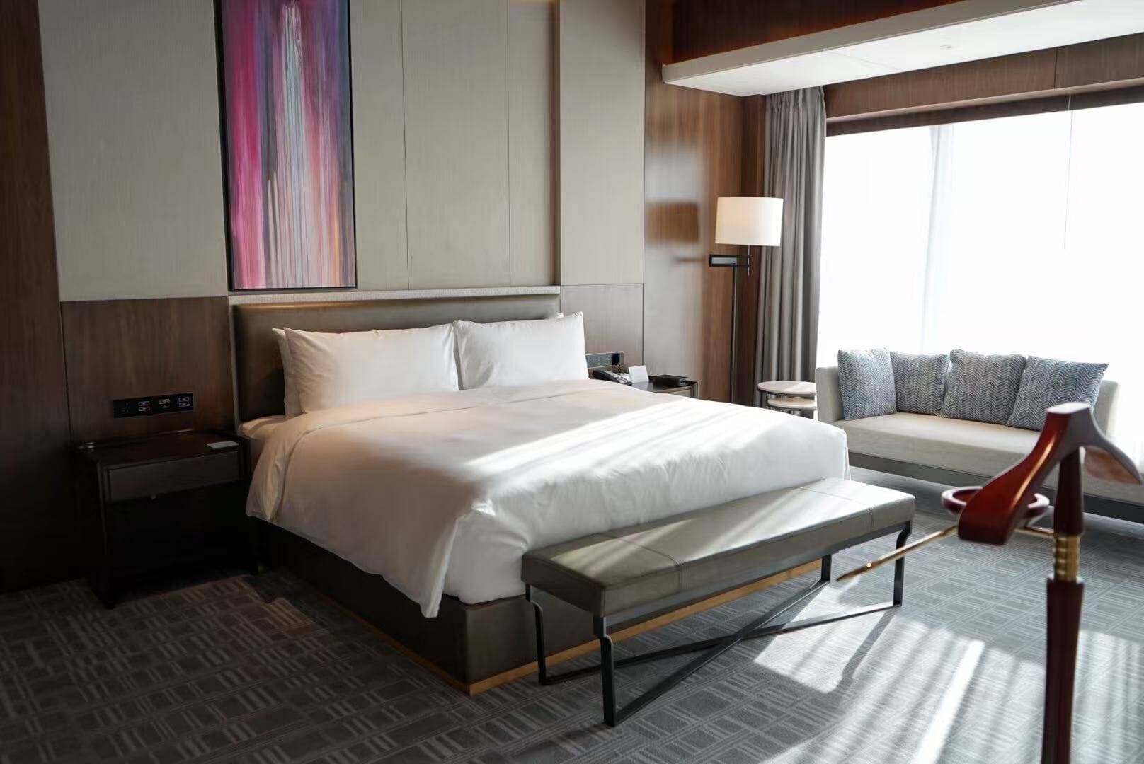 A hotel room with a bed, bedside tables, a desk, a lamp, a bench, and a sofa with cushions.