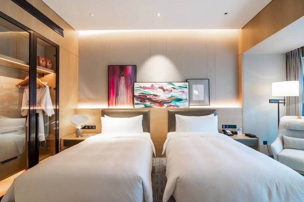 A hotel room with two beds, a wardrobe, a desk, a lamp, and artwork on the wall.